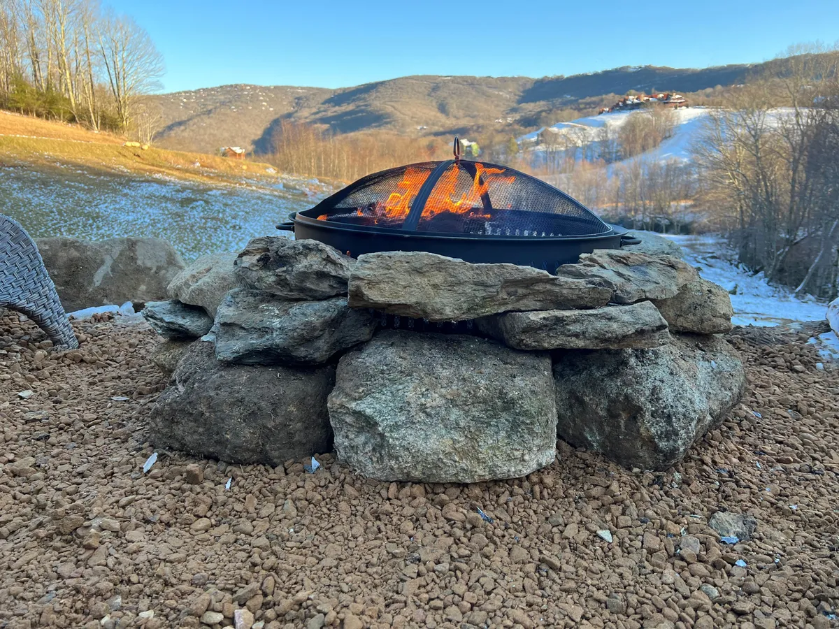 A cozy outdoor fire pit encircled by natural rock seating, set against a backdrop of distant mountains. The scene evokes togetherness as families gather around the warm glow, roasting marshmallows or sharing stories beneath the night sky. The environment is inviting, peaceful, and perfect for winding down after a day of adventure.