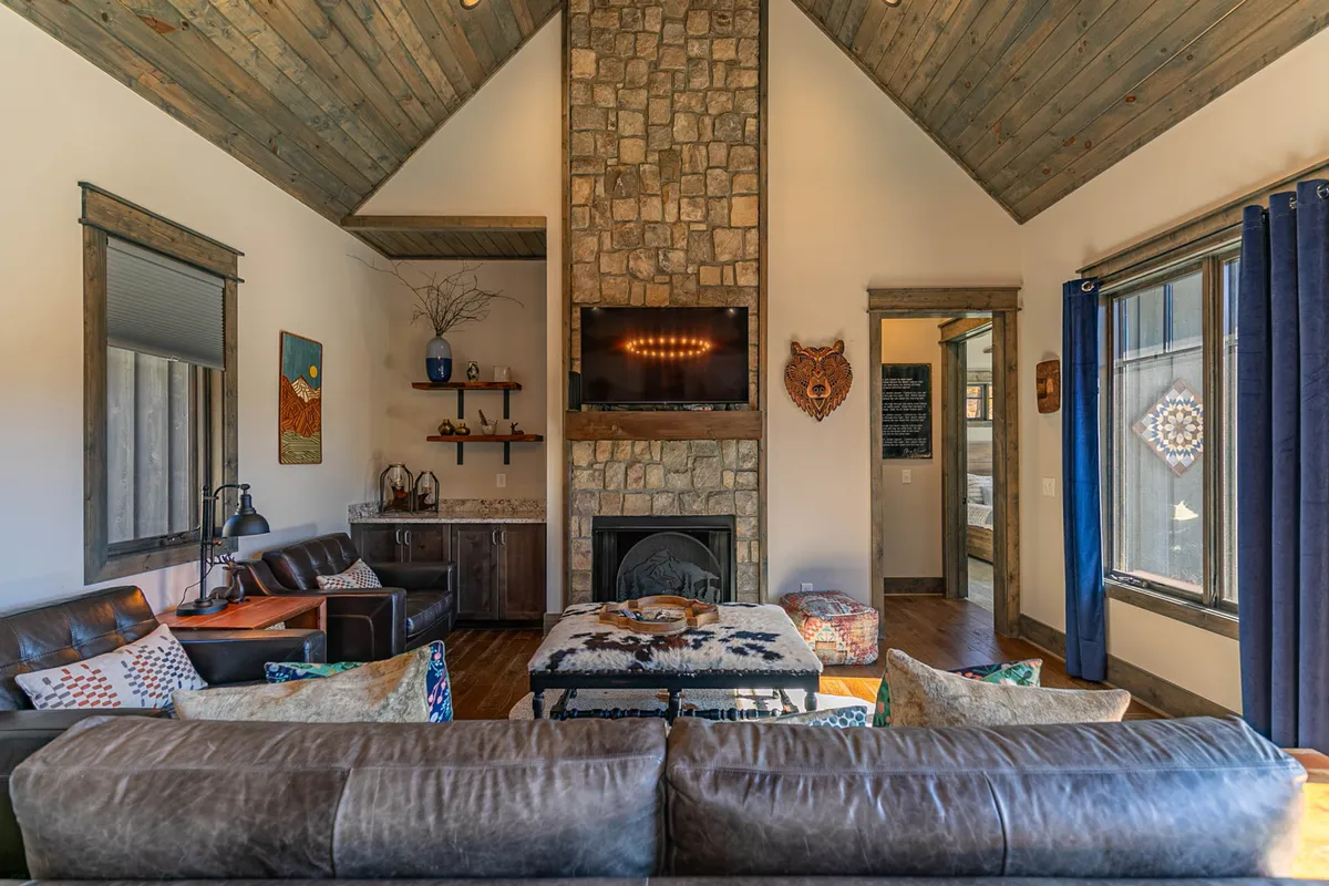 A warmly lit living area with a stone fireplace, large windows letting in mountain views, cozy sofas, and decorative wall art. The space radiates a welcoming atmosphere ideal for socializing, relaxing, or reading by the fire.