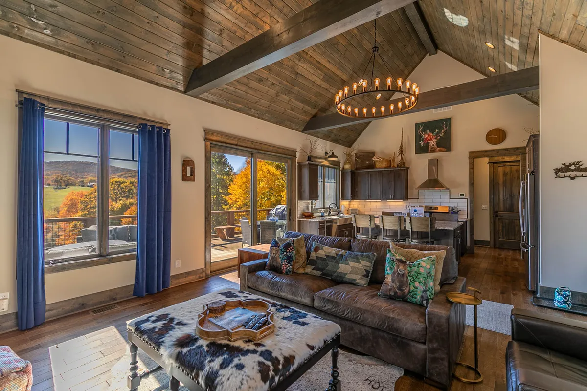 A stylish living room with a comfortable sofa adorned with patterned cushions, a chandelier overhead, large windows with natural light, exposed wooden beams, and a coffee table. The space feels warm, bright, and ready for gatherings or quiet relaxation.