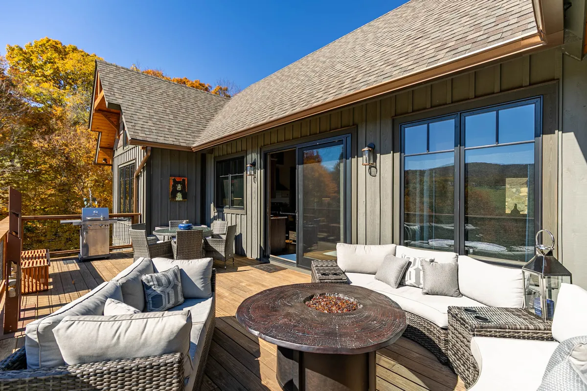 An inviting outdoor seating area featuring a modern fire pit table surrounded by comfortable chairs, a grill set up nearby, large windows reflecting autumn foliage, and a soft glow of evening light over the wooded landscape.