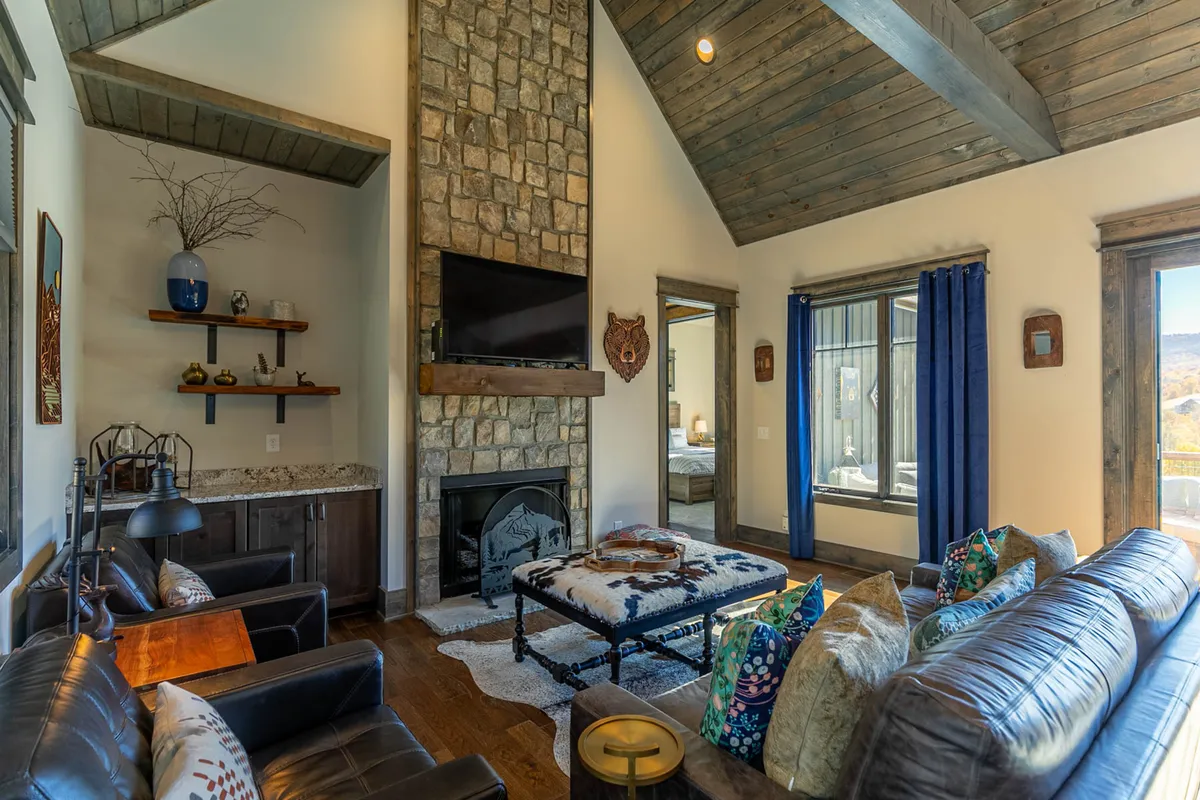 A cozy living area featuring a stone fireplace, comfortable seating with colorful cushions, and large windows that display beautiful outdoor views. The shelves are decorated, and the firelight suggests a welcoming spot to unwind after a day of adventure.