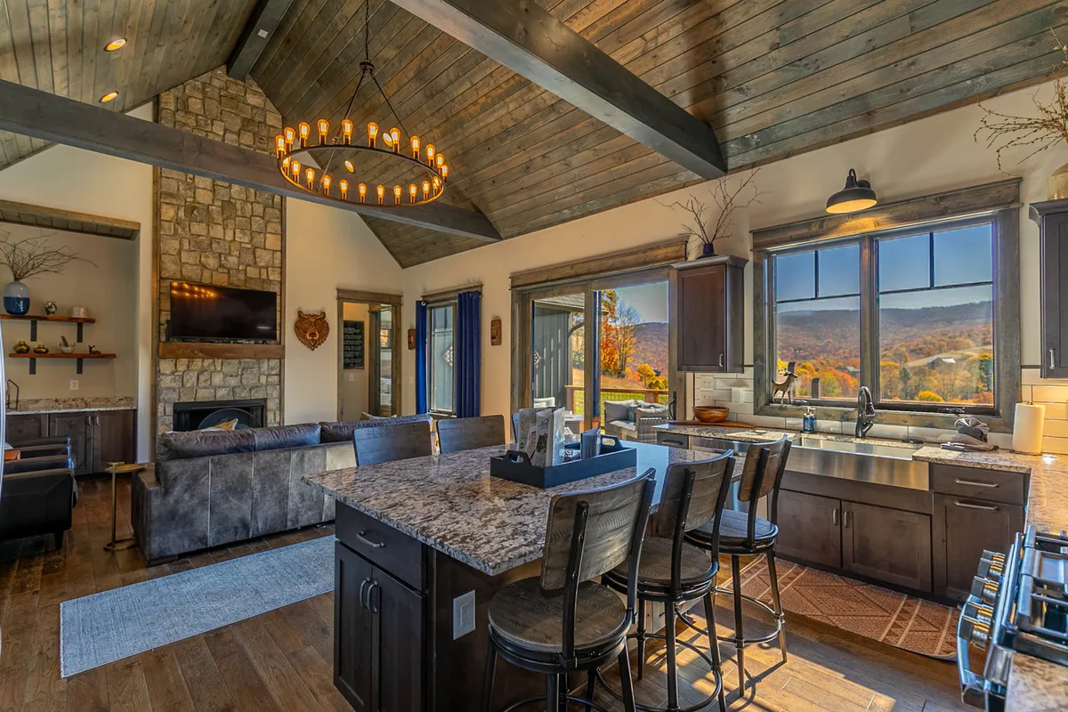A spacious kitchen with a large island, elegant chandelier, and a prominent stone fireplace. Floor-to-ceiling windows frame panoramic views of the surrounding mountains, creating a seamless connection between indoors and the outdoors. The scene evokes a sense of celebration, gathering, and taking in the mountain scenery.