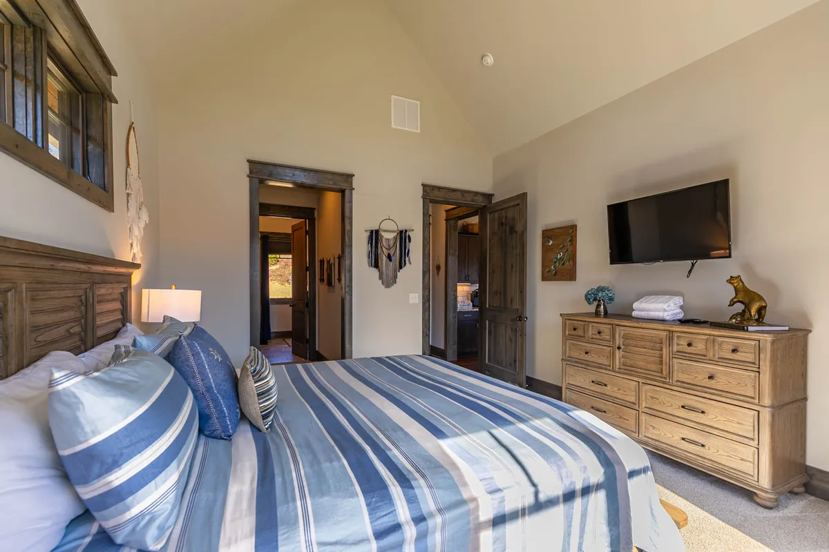 A cozy bedroom with a striped bedspread, wooden dresser, wall-mounted TV, and decorative wall hangings, filled with natural light. The space feels inviting and modern, catering to both comfort and entertainment.
