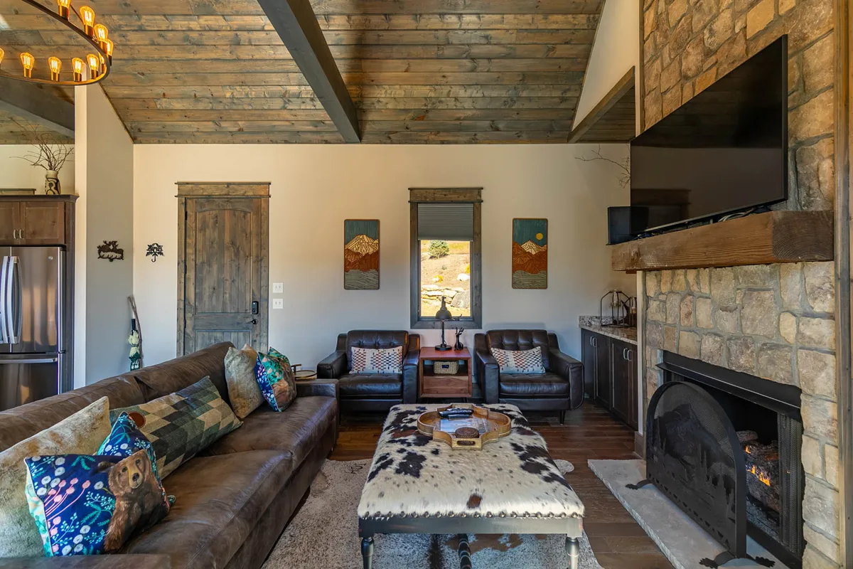 A spacious great room featuring a large plush leather sofa, a prominent stone fireplace, wooden beams overhead, mountain-themed artwork, and a cowhide rug. Warm natural light fills the room, highlighting the cozy and inviting atmosphere.