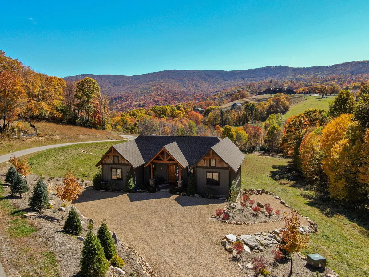 The image showcases a beautiful single-story mountain home set against a backdrop of rolling hills and vibrant, colorful trees. The house features a spacious outdoor area perfect for gatherings, with sweeping views of the Blue Ridge Mountains. A cozy fire pit and inviting seating hint at relaxing evenings, while the surrounding landscape invites guests to explore nature trails or simply take in the sunset.