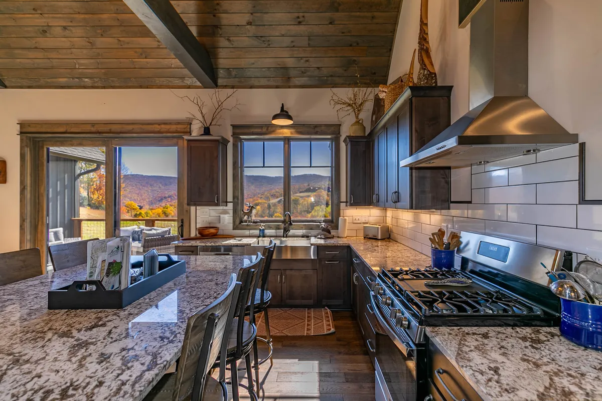 A warmly lit kitchen space with a granite countertop, large window offering mountain views, exposed wooden beams, and gleaming stainless-steel appliances. The scene feels cozy and invites guests to gather for coffee or casual meals while enjoying scenic views.
