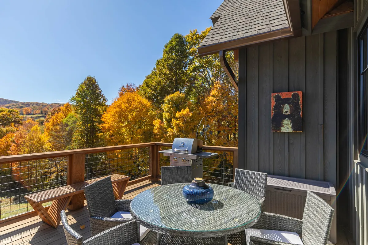 An outdoor dining area on a deck surrounded by trees, featuring a glass table, grill, and whimsical bear art. The tranquil woods create a peaceful setting for shared meals, while the setup suggests relaxed gatherings, barbecuing, and time spent savoring the mountain air with family and pets.